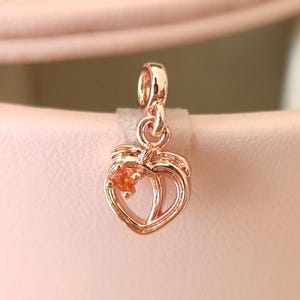 May include: Rose gold peach charm with a small orange gemstone. The charm has a detailed design with a loop at the top for attaching to a bracelet or necklace. The peach charm is a pendant.