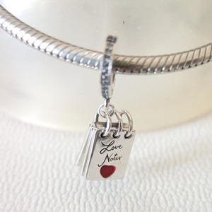 May include: A silver charm with a small notepad design. The notepad has the words "Love Notes" written on it, with a red heart at the bottom. The charm is attached to a silver bracelet.