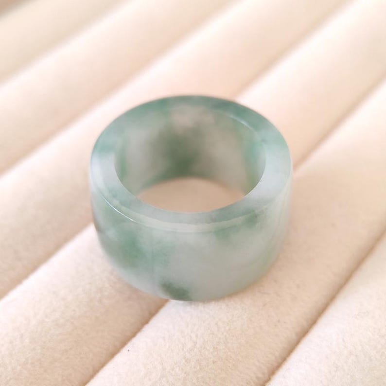 K&ouml;nnte beinhalten: Ein breiter, runder Ring aus Jade. Der Ring hat eine hellgr&uuml;ne Farbe mit wei&szlig;en Marmorierungen. Der Ring ist glatt und poliert. Der Ring scheint ein massives St&uuml;ck Jade zu sein.