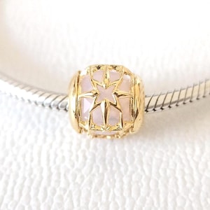 May include: A gold-colored charm with a star design and a light pink center. The charm is cylindrical with a starburst pattern on the front and is attached to a silver bracelet. The charm is likely a jewelry accessory.