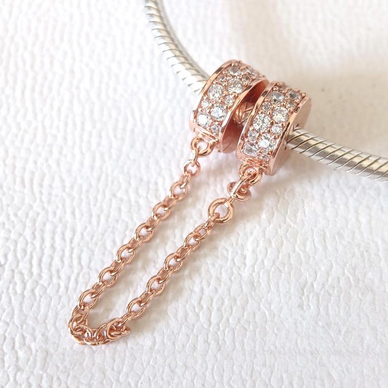 May include: Rose gold charm with two ring-shaped beads encrusted with small, clear stones. A delicate chain connects the two beads. The charm is displayed on a silver bracelet.