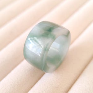 K&ouml;nnte beinhalten: Ein breiter, transluzenter Jade-Ring mit glatter, polierter Oberfl&auml;che. Der Ring weist eine Mischung aus hellgr&uuml;nen und wei&szlig;en Farbt&ouml;nen auf, wodurch ein Marmoreffekt entsteht. Das Design des Rings ist schlicht und elegant, f&uuml;r den Alltag geeignet.