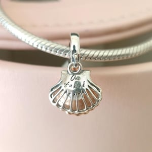 Op de afbeelding: Een zilveren schelpenbedel met een parelachtig midden, hangend aan een zilveren ketting. De bedel heeft de tekst "One of a Kind" gegraveerd. De achtergrond is zachtroze.