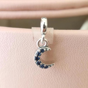 Könnte beinhalten: Ein silberfarbener Anhänger mit einem Halbmond-Design. Der Mond ist mit kleinen, dunkelblauen Edelsteinen besetzt. Der Anhänger hängt an einer kleinen Schlaufe, vor einem zartrosa Hintergrund.