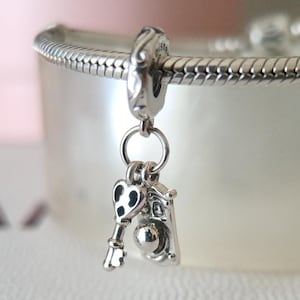 Könnte beinhalten: Ein silberner Charm mit einem Schlüssel- und Kamera-Design. Der Schlüssel hat einen herzförmigen Kopf mit schwarzen Akzenten. Der Kamera-Charm hat eine runde Linse und einen rechteckigen Körper. Der Charm ist an einem Silberarmband befestigt.