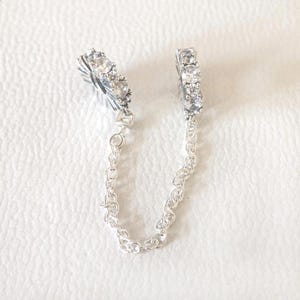 Può includere: Catena di sicurezza argentata per bracciale con ciondoli. La catena presenta due estremità decorative, ciascuna con pietre chiare e scintillanti. Una sottile catena collega le due estremità, offrendo sicurezza ed eleganza.