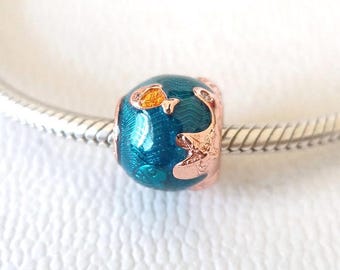 Puede incluir: Una cuenta de dije de color turquesa y oro rosa con un diseño de globo terráqueo. La cuenta presenta un acabado de esmalte turquesa con detalles en oro rosa que representan continentes y detalles brillantes. El dije está en una pulsera plateada.