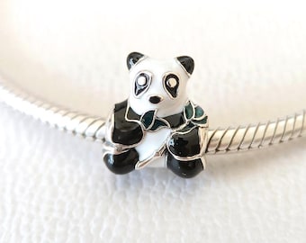 May include: A panda bear charm with black and white enamel details. The charm has black ears, eyes, and feet, with a white face and body. The panda is holding a green leaf. The charm is on a silver bracelet.