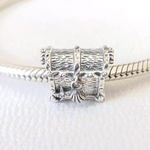 Puede incluir: Un charm de plata con forma de cofre del tesoro. El charm es muy detallado con vetas de madera texturizadas y detalles metálicos decorativos. Una pequeña borla cuelga de la parte delantera. El charm está en una pulsera de plata.