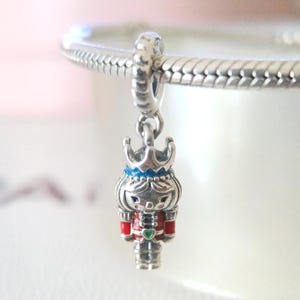 Pode incluir: Um berloque de prata com um soldado quebra-nozes em miniatura com uma coroa. O berloque tem um uniforme vermelho e preto com um detalhe verde em forma de coração. A coroa do quebra-nozes é azul e prateada.