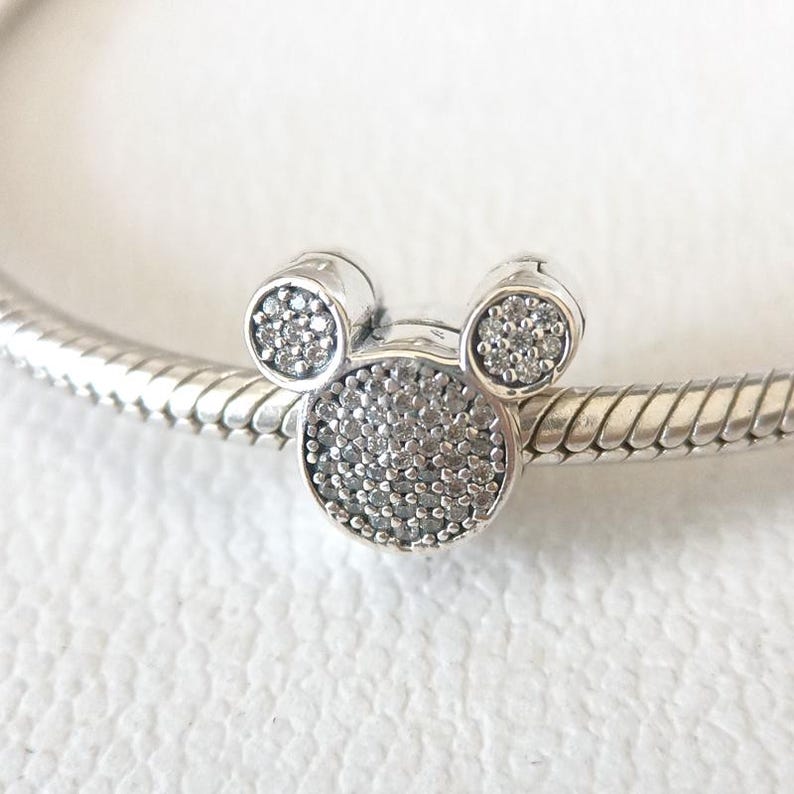 Op de afbeelding: Een zilveren bedel in de vorm van Mickey Mouses hoofd, met twee kleine ronde oren en een groter rond hoofd. De bedel is bezet met kleine, sprankelende, heldere steentjes. De bedel zit aan een zilveren armband.