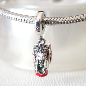 May include: A silver charm depicting a miniature figure with an ornate headdress and red detailing. The charm hangs from a silver chain, displaying a design with pink, red, and blue gemstones.