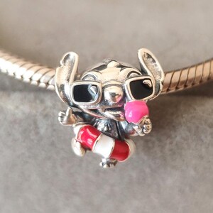 K&ouml;nnte beinhalten: Ein silberner Charm-Anh&auml;nger, der die Figur Stitch mit schwarzer Sonnenbrille zeigt, die eine rosa Eist&uuml;te h&auml;lt und einen rot-wei&szlig; gestreiften Rettungsring tr&auml;gt. Der Charm ist auf ein silbernes Schlangenketten-Armband aufgef&auml;delt.