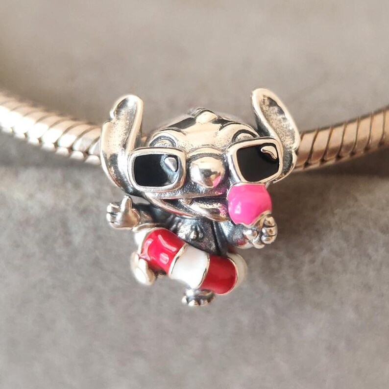 K&ouml;nnte beinhalten: Ein silberner Charm-Anh&auml;nger, der die Figur Stitch mit schwarzer Sonnenbrille zeigt, die eine rosa Eist&uuml;te h&auml;lt und einen rot-wei&szlig; gestreiften Rettungsring tr&auml;gt. Der Charm ist auf ein silbernes Schlangenketten-Armband aufgef&auml;delt.