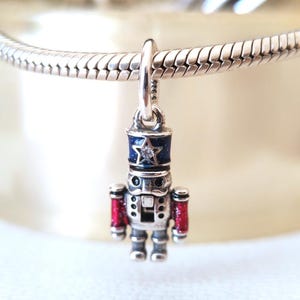 May include: A silver charm of a toy soldier nutcracker. The nutcracker has a blue hat with a star, red arms, and a silver body. The charm is suspended from a silver bracelet.