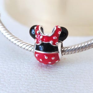 May include: A charm featuring a red and black design, reminiscent of a cartoon character's outfit. The charm has a red top with white polka dots, a black midsection, and black circular accents. It is attached to a silver bracelet.