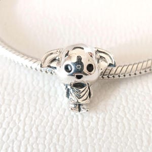May include: A silver charm depicting a stylized character with large ears, large eyes, and a small robe. The charm is attached to a silver chain bracelet. The charm is highly detailed and has a polished finish.