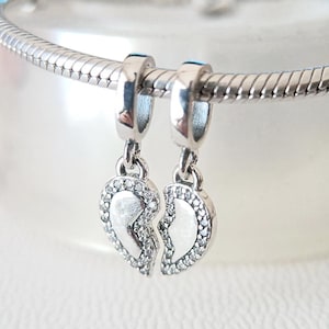 Könnte beinhalten: Ein silbernes Charm-Armband mit zwei herzförmigen Anhängern. Jeder Anhänger ist geteilt, wobei eine Seite mit kleinen, klaren Steinen besetzt ist. Die Anhänger sind mit silbernen Schlaufen am Armband befestigt.