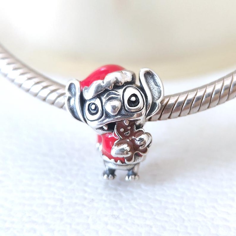 May include: A silver charm featuring a cartoon character wearing a red Santa hat and coat, holding a gingerbread man. The charm is detailed with black eyes and is attached to a silver bracelet.