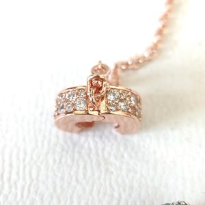 May include: Rose gold necklace pendant with a row of small, clear gemstones. The pendant is a rounded shape with a clasp. The chain is also rose gold and has a twisted rope design. The background is white.