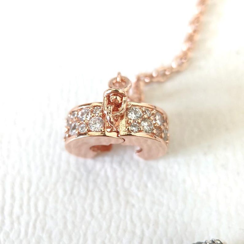 May include: Rose gold necklace pendant with a row of small, clear gemstones. The pendant is a rounded shape with a clasp. The chain is also rose gold and has a twisted rope design. The background is white.