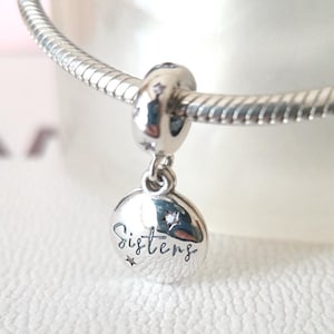 Può includere: Un charm in argento con la scritta "Sisters" in corsivo, impreziosito da piccole stelle. Il charm è appeso a un braccialetto d'argento. Il charm rotondo ha una piccola gemma.