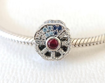 May include: A silver charm with a wheel design, featuring a central round red gemstone. The wheel is detailed with clear and blue gemstones. The charm is attached to a silver bracelet.