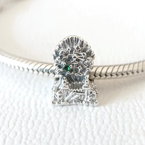 Puede incluir: Un charm de plata con un diseño detallado de un trono, con una pequeña gema verde. El charm está unido a una pulsera de plata. El charm tiene detalles intrincados y un estilo vintage.