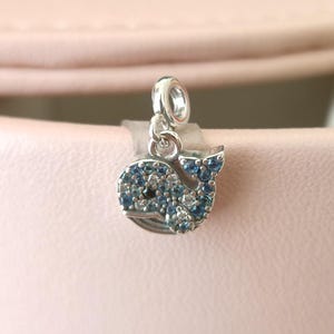 Może przedstawiać: Srebrny charms w kształcie wieloryba, inkrustowany niebieskimi i przezroczystymi kryształami. Charms jest przeznaczony do mocowania do bransoletki lub naszyjnika. Ciało wieloryba jest szczegółowo wykonane z mieszanki jasnych i ciemnoniebieskich kryształów.