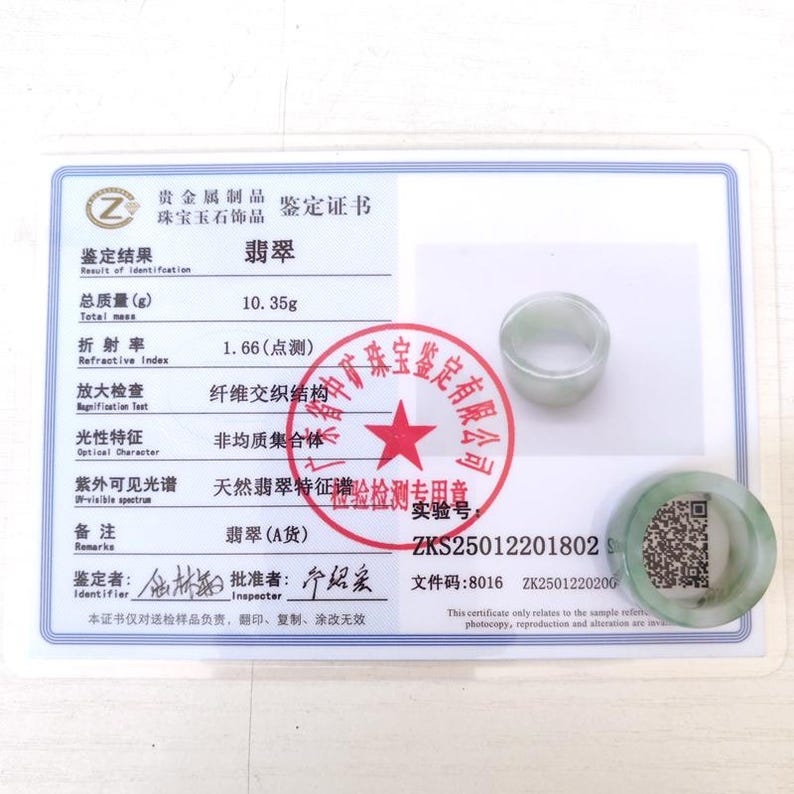 K&ouml;nnte beinhalten: Ein Jade-Ring und eine Jade-Scheibe liegen auf einem Zertifikat. Das Zertifikat enth&auml;lt chinesischen Text und Details zu den Eigenschaften des Jades, einschlie&szlig;lich Gewicht (10,35 g) und Brechungsindex (1,66). Ein roter Stempel mit chinesischem Text ist ebenfalls sichtbar.