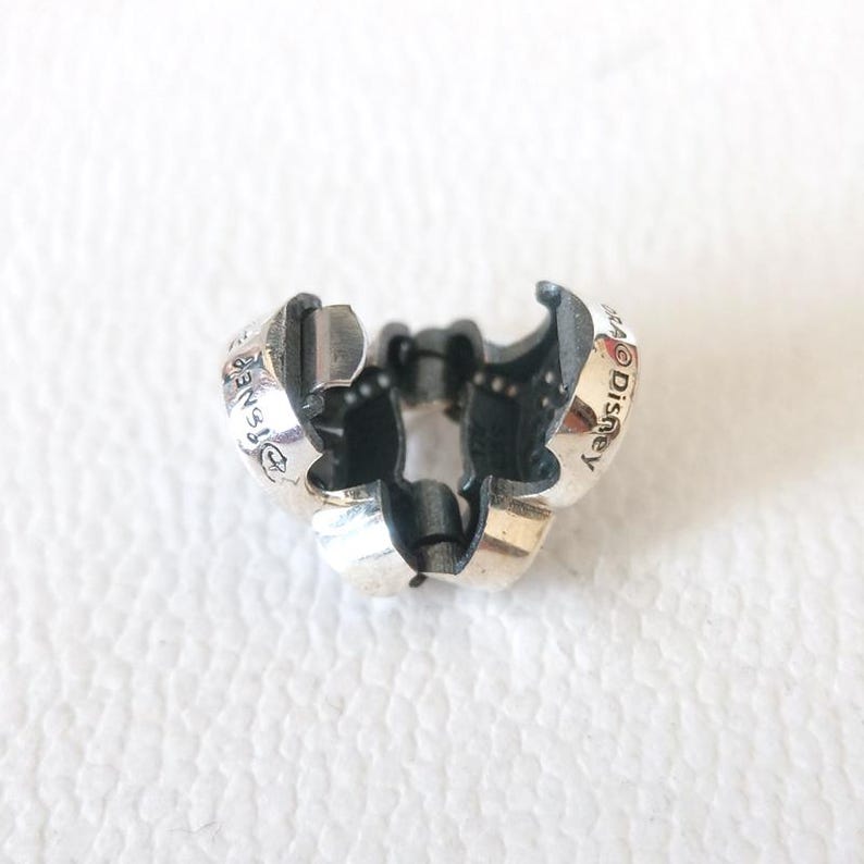 Op de afbeelding: Een zilverkleurige bedel met een driedelig ontwerp. De bedel heeft het woord "Disney" op het oppervlak gegraveerd. De bedel is ontworpen als sluiting voor een armband of ketting.