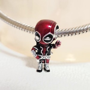 Puede incluir: Un charm de Deadpool en rojo, negro y plateado. El charm es una figura en miniatura del personaje de Marvel Comics, con una máscara roja, un traje negro y detalles plateados. El charm está unido a una pulsera plateada.