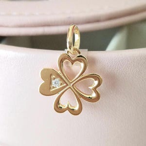 May include: Gold-colored clover pendant with four heart-shaped leaves. A small, clear gemstone is set in the center. The pendant is suspended from a loop at the top, ready to be added to a necklace or bracelet.