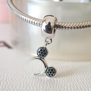 May include: A silver charm with a Mickey Mouse design, featuring two black-studded circles and a curved shape. The charm is attached to a silver bracelet, showcasing a polished, detailed design.