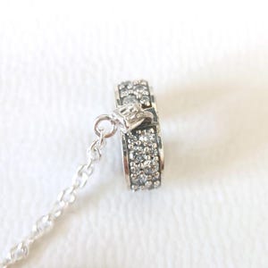 May include: A silver charm with a ring design, encrusted with small, clear gemstones. The charm is attached to a delicate silver chain. The ring charm is approximately 0.5 inches in diameter.
