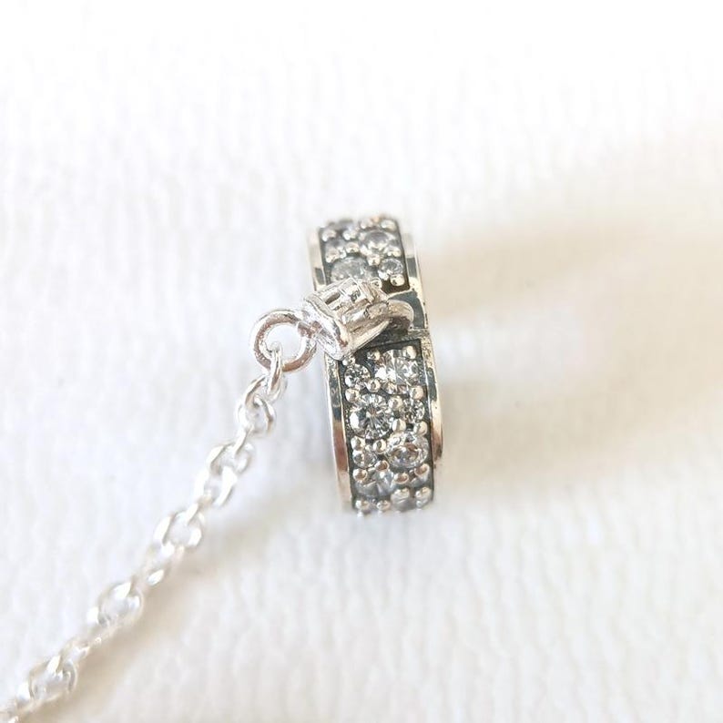May include: A silver charm with a ring design, encrusted with small, clear gemstones. The charm is attached to a delicate silver chain. The ring charm is approximately 0.5 inches in diameter.