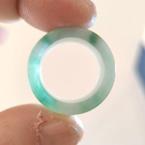 K&ouml;nnte beinhalten: Ein transluzenter Jade-Ring mit einem lebendigen Gr&uuml;nton. Der Ring wird zwischen zwei Fingern gehalten und zeigt seine polierte Oberfl&auml;che und sein kreisf&ouml;rmiges Design. Der Jade weist nat&uuml;rliche Farbvariationen auf, was seinen einzigartigen Reiz unterstreicht. Durchmesser: ca. 2 cm.