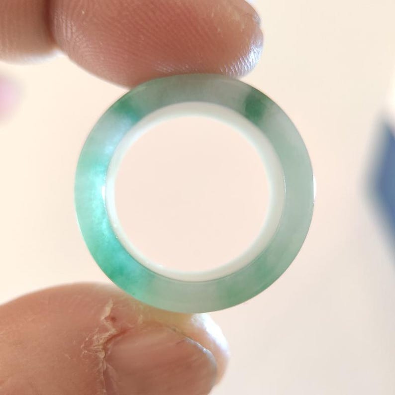 K&ouml;nnte beinhalten: Ein transluzenter Jade-Ring mit einem lebendigen Gr&uuml;nton. Der Ring wird zwischen zwei Fingern gehalten und zeigt seine polierte Oberfl&auml;che und sein kreisf&ouml;rmiges Design. Der Jade weist nat&uuml;rliche Farbvariationen auf, was seinen einzigartigen Reiz unterstreicht. Durchmesser: ca. 2 cm.