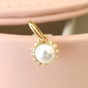 Könnte beinhalten: Ein goldfarbener Anhänger mit einem weißen Perlenmittelpunkt, umgeben von einem Sonnenstrahlendesign. Der Anhänger ist an einem kleinen goldenen Reifen befestigt. Der Hintergrund ist rosa.