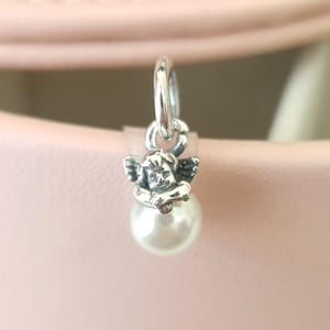 Puede incluir: Un charm de plata con un querubín con alas detalladas y una perla. El charm cuelga de un aro de plata. El fondo es rosa pálido, creando un delicado contraste con la plata.