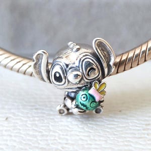 Puede incluir: Un charm de plata con el personaje Stitch de la pel&iacute;cula Lilo & Stitch. Stitch sostiene un objeto redondo y colorido con un cuerpo verde, un lazo rosa y alas amarillas. El charm es detallado y parece ser de metal.