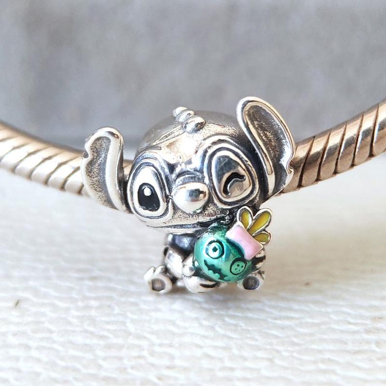 Puede incluir: Un charm de plata con el personaje Stitch de la pel&iacute;cula Lilo & Stitch. Stitch sostiene un objeto redondo y colorido con un cuerpo verde, un lazo rosa y alas amarillas. El charm es detallado y parece ser de metal.