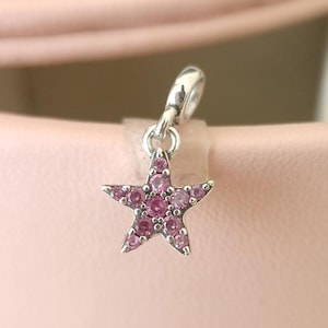 May include: A silver charm with a star design. The star is filled with small, round, pink gemstones. The charm has a loop at the top for attaching to a bracelet or necklace. The background is a soft pink.