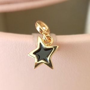 May include: A gold-colored star charm with a black enamel center. The charm has a loop at the top for attaching to a bracelet or necklace. The star is a classic shape, and the gold and black color combination gives it a modern look.