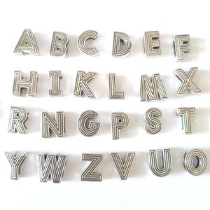 May include: Silver-toned alphabet charms, each letter from A to Z, displayed on a white surface. The charms have a layered design, with each letter clearly defined. Ideal for jewelry making or crafting projects.