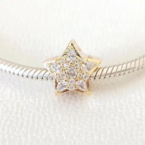 Puede incluir: Un charm de estrella dorada con un grupo de piedras transparentes y brillantes en el centro. La estrella está en una pulsera plateada texturizada. El charm es una pieza de joyería.