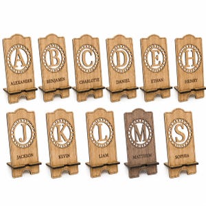 Puede incluir: Una colecci&oacute;n de once soportes de madera para tel&eacute;fonos, cada uno con un recorte circular con una letra may&uacute;scula cortada a l&aacute;ser y un nombre grabado debajo. Los soportes tienen una base con muescas y un acabado de madera clara.