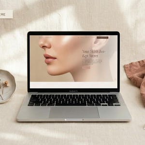 May include: A silver MacBook laptop displaying a website with the text "Your Skin's Best-Kept Secret" and a side profile of a face. A small ceramic dish with dried plants and a folded cloth are next to the laptop. A tag reads "Skin Secret Aesthetics".