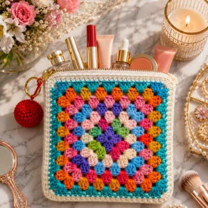 Puede incluir: Un neceser cuadrado de ganchillo con un diseño de granny square colorido. La bolsa contiene varios artículos de maquillaje, como barras de labios y tubos. Un llavero de pompón rojo cuelga de la cremallera.