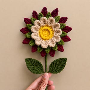 Puede incluir: Una flor de ganchillo hecha a mano con un centro amarillo brillante, un anillo de pétalos beige claro y capas de pétalos puntiagudos en color burdeos oscuro y verde bosque. La flor está unida a un tallo verde fino con dos hojas texturizadas.